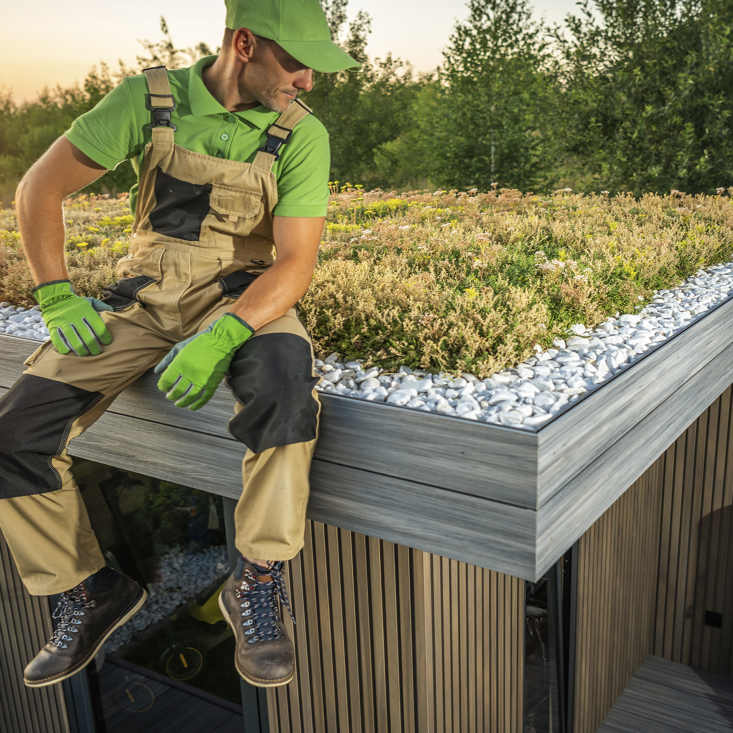 A handyman in green gloves and work attire sitting on an eco-friendly rooftop garden with lush vegetation and white pebbles.